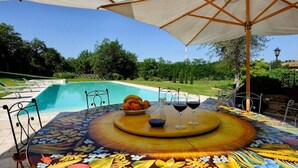 A dining table with a floral tablecloth, surrounded by outdoor seating, a pool, and a view of a landscaped garden.