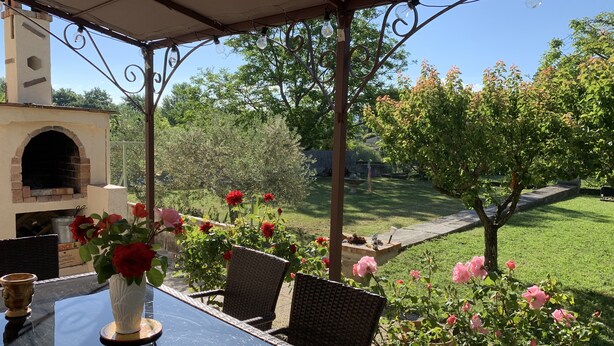 Un porche cubierto con una mesa de vidrio y sillas de mimbre, con vistas a un jardín con rosales y un horno de ladrillo.