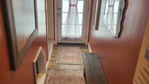 A hallway with a wooden bench, a rug, a door with sheer curtains, and a wall sign that reads "IN GOD WE TRUST". - Cozy Guest House on 15 acres, with plenty of parking! (Parsonsburg)