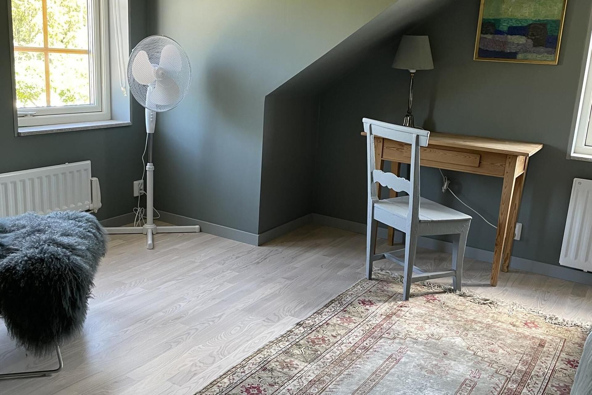 Una stanza con una scrivania in legno, una sedia bianca, una ventola portatile, un termosifone, una poltrona soffice e un tappeto decorato.