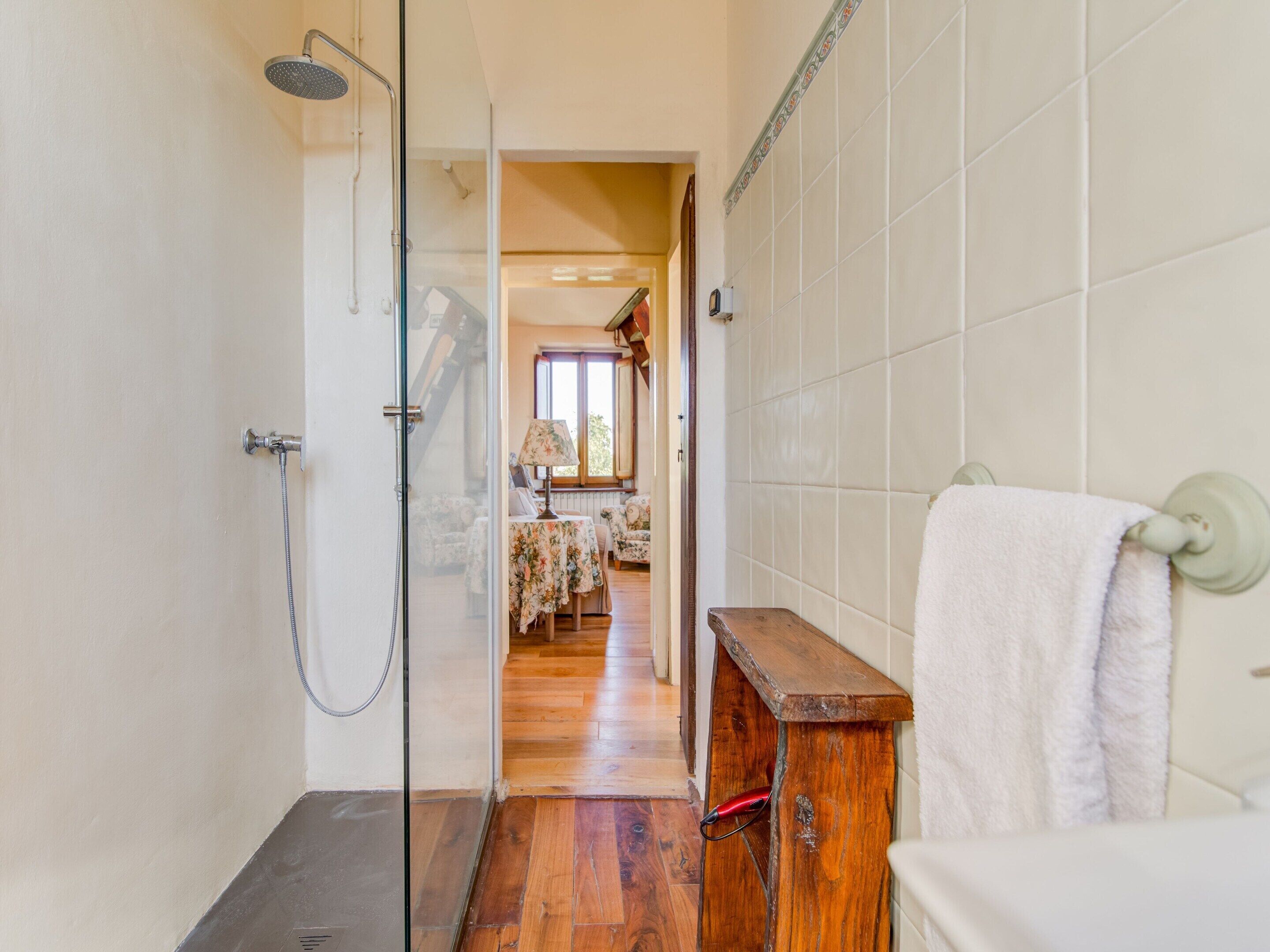 A bathroom with a glass shower enclosure, a wooden bench, and a white towel hanging on the wall.