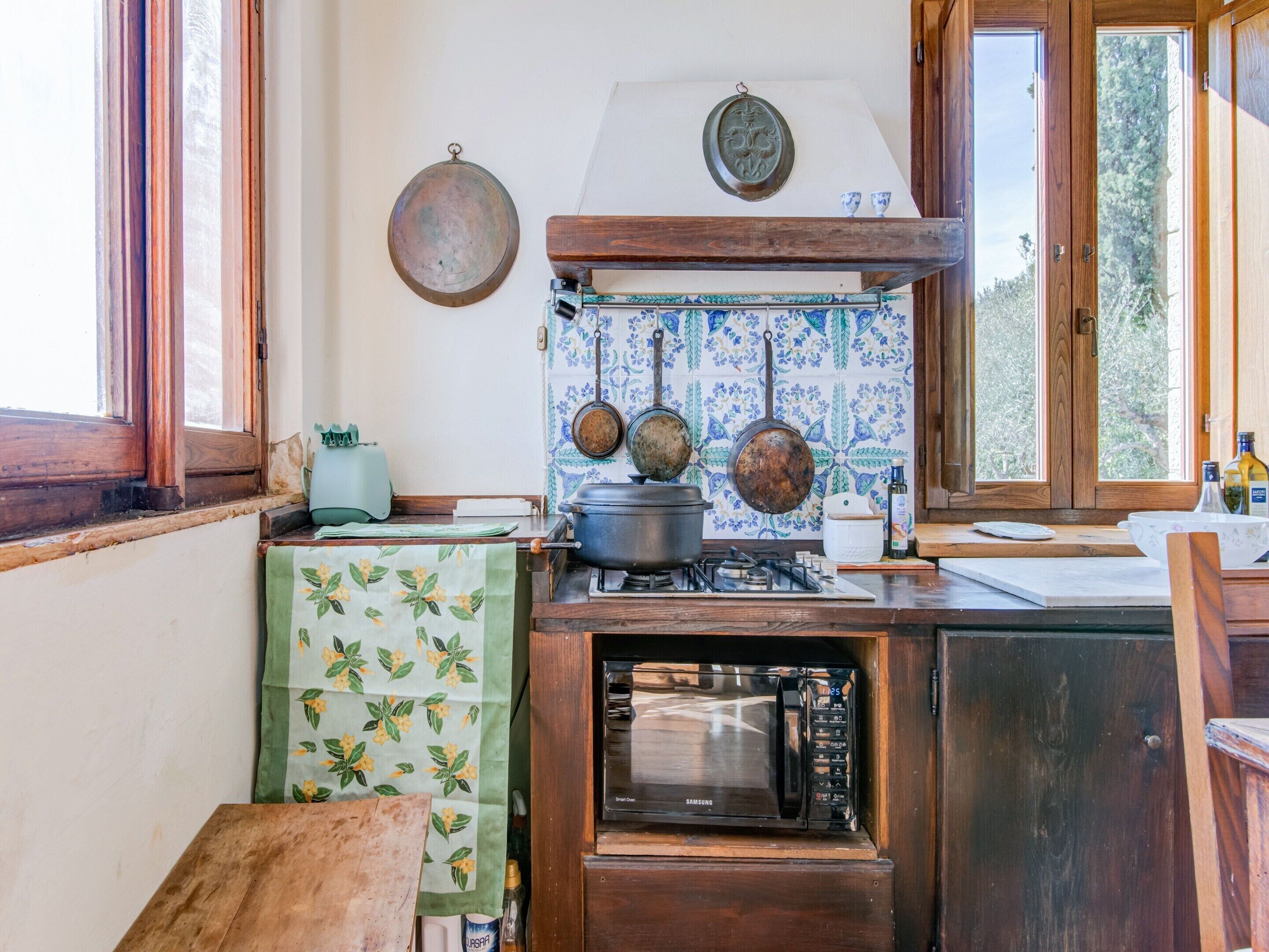 A kitchen with a stove, microwave, hanging pots, and a window with a view of greenery.