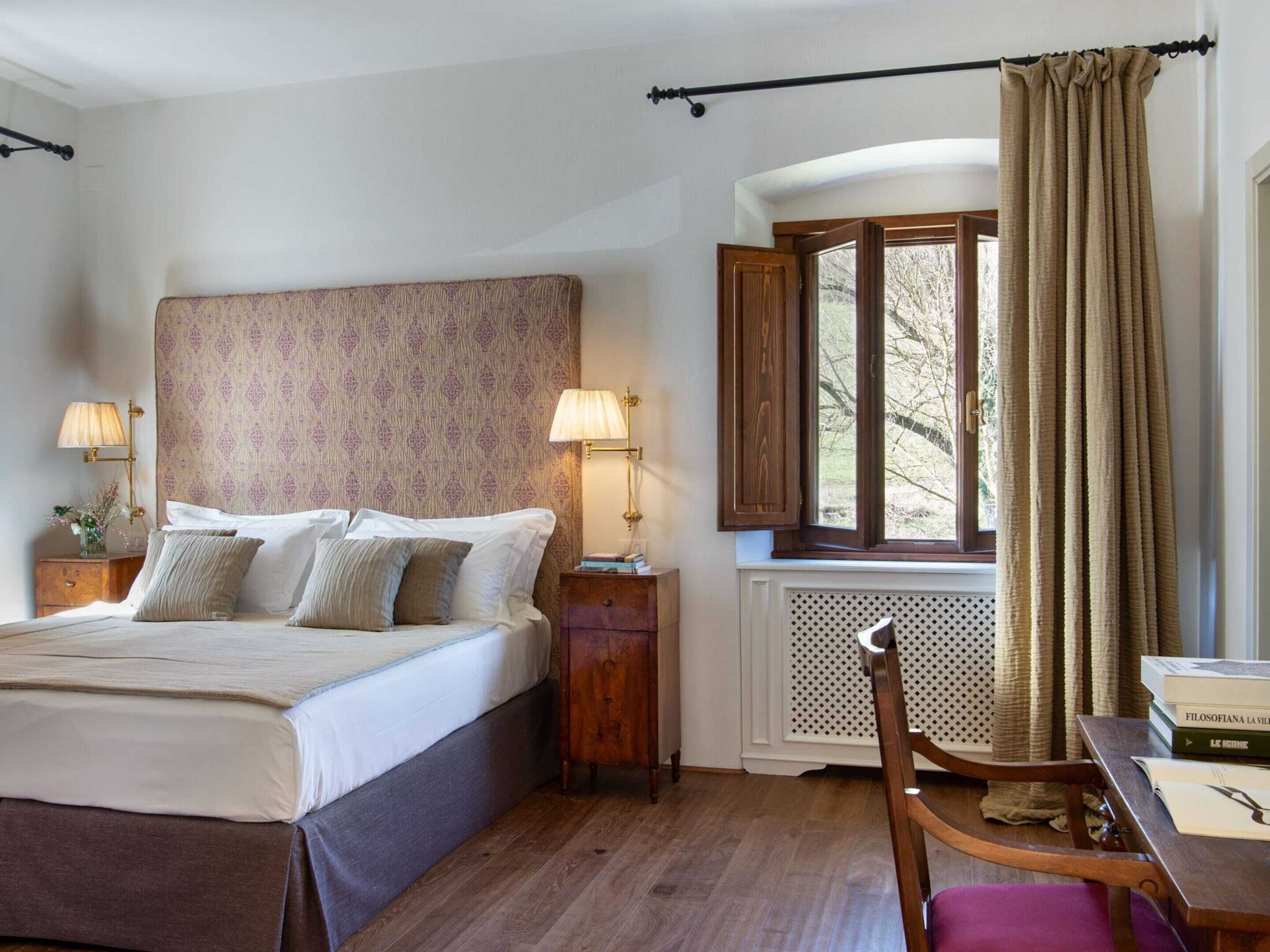 A neatly arranged bedroom with a bed, bedside lamps, a window with curtains, a radiator, and a wooden chair.