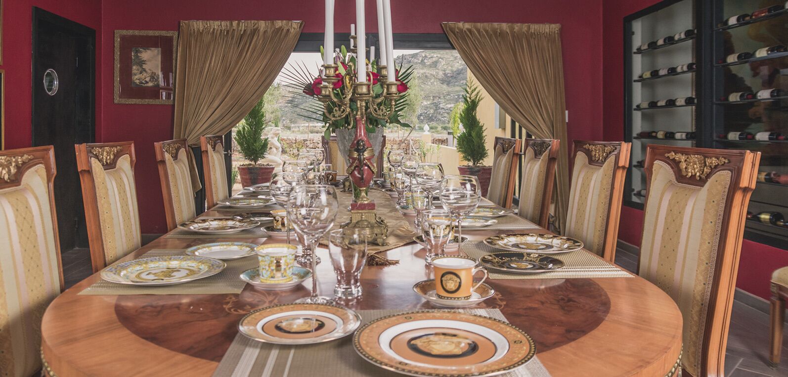 A formal dining room with a long table set for a meal, flanked by ornate chairs, and a wine rack in the background.