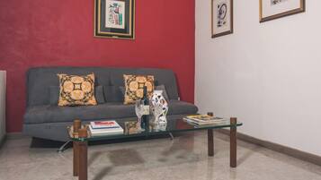Ein Wohnzimmer mit grauem Sofa, zwei gemusterten Kissen, einem Glastisch mit Büchern und einer Vase sowie gerahmten Kunstwerken an der roten Wand.