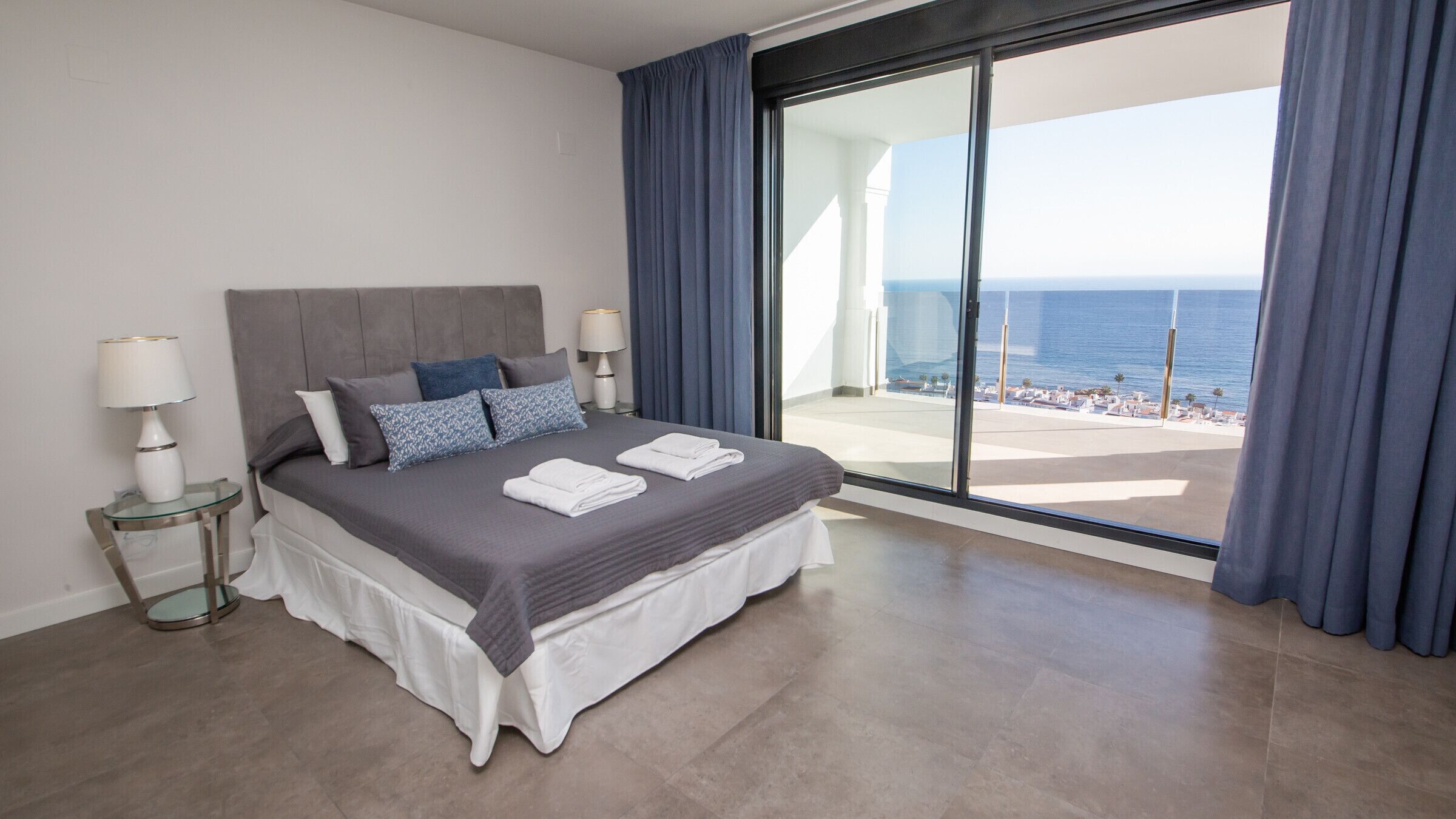 A modern hotel room with a large bed, a glass door leading to a balcony with a view of the sea.