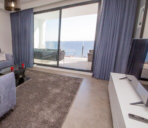 A modern living room with a grey sofa, a glass coffee table, and a large window offering a view of the sea.