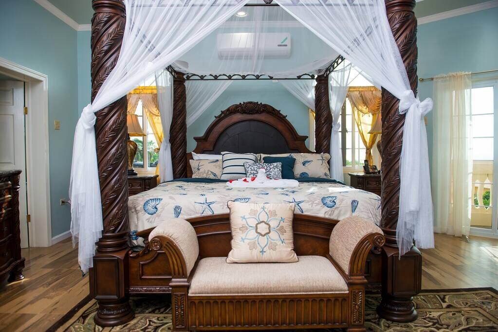 A four-poster canopy bed with a floral bedspread, a wooden bench, and a nightstand with a lamp.