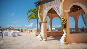 Espaço para casamento ao ar livre com cortinas brancas e douradas, uma palmeira e vista para o oceano.