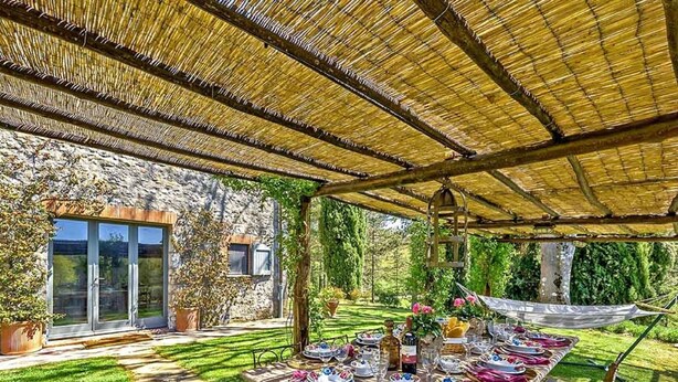 Un'area pranzo all'aperto con un tavolo apparecchiato per un pasto, circondata da un edificio in pietra e un giardino.
