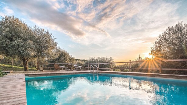 Una piscina con muelle de madera, rodeada de árboles y con un atardecer de fondo.