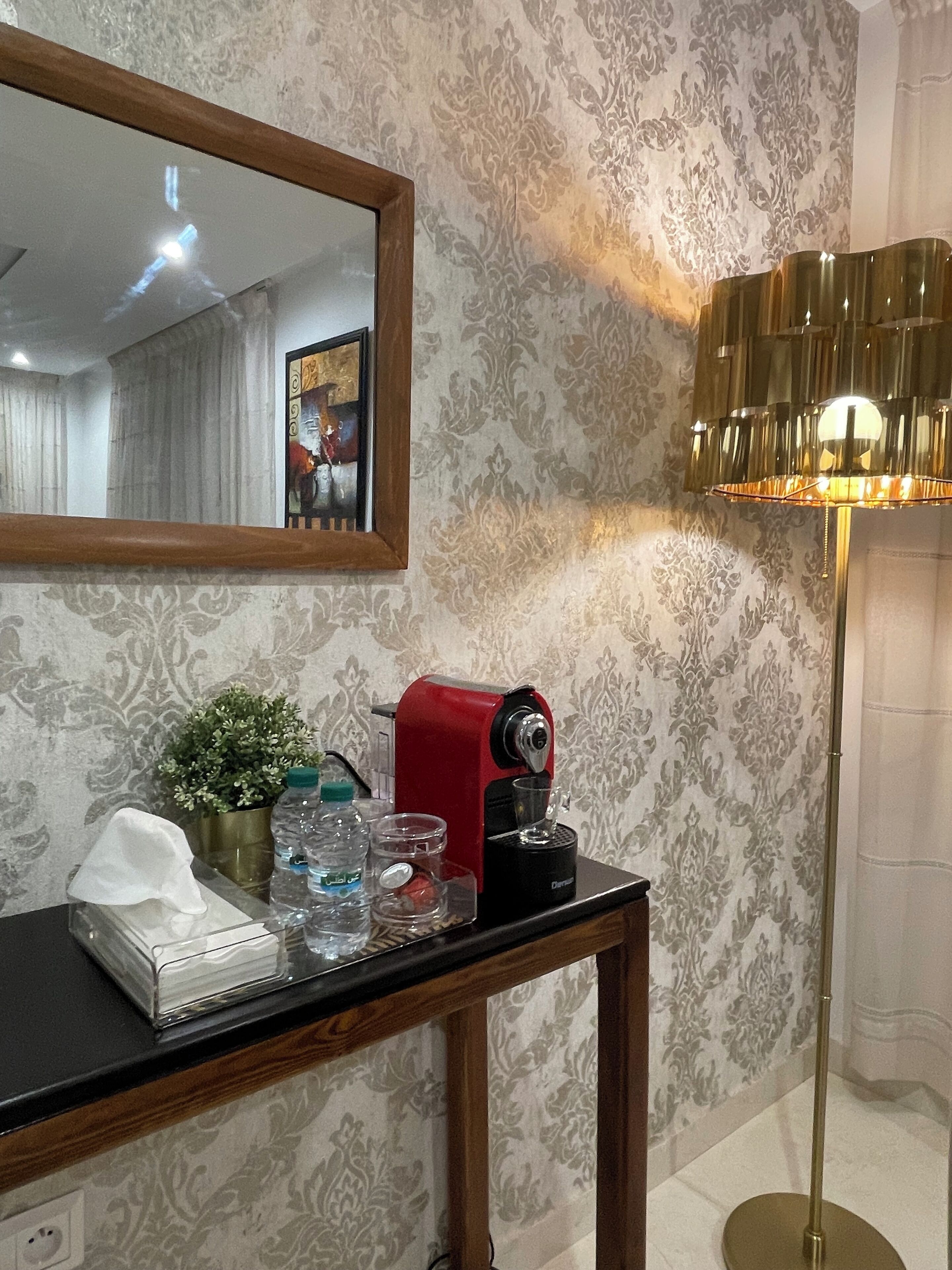 Una habitación con una mesa de madera, una cafetera roja, una planta en maceta, un espejo y una lámpara de pie.