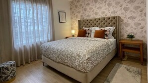 Une chambre à coucher avec un grand lit, une table de chevet, une petite table, une plante en pot et une fenêtre avec des rideaux.