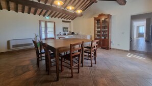 A spacious dining area with a large wooden table and chairs, a kitchen in the background, and a wooden cabinet with glassware. - Tasteful Countryside Stay (Montalcino)