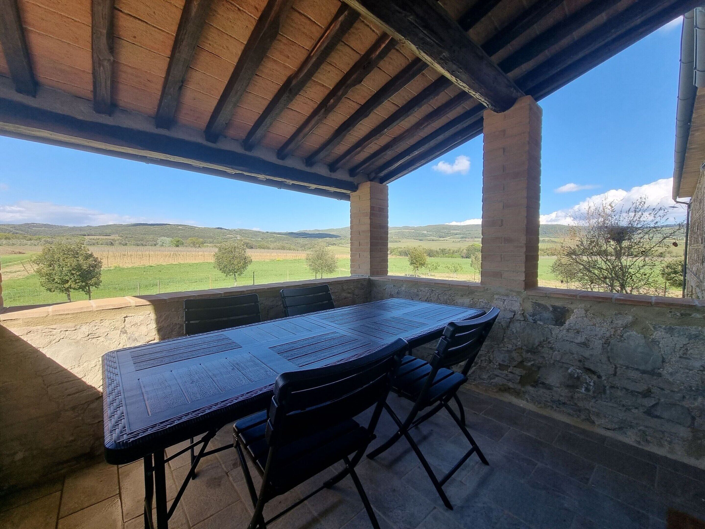 Uma área de refeições coberta ao ar livre, com mesa e cadeiras, paredes de pedra e vista para uma paisagem verdejante.