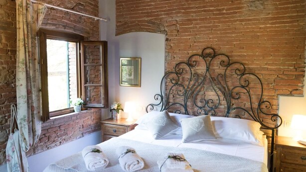 A bedroom with a brick wall, a bed with white linens, a decorative metal headboard, and a window with curtains. - Farmhouse in Tuscany With Pool & Near Montecastello (Pontedera)