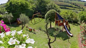 Un parque infantil con columpios, un tobogán y una pequeña estructura de madera en un área con césped, árboles y vistas a colinas ondulantes al fondo.