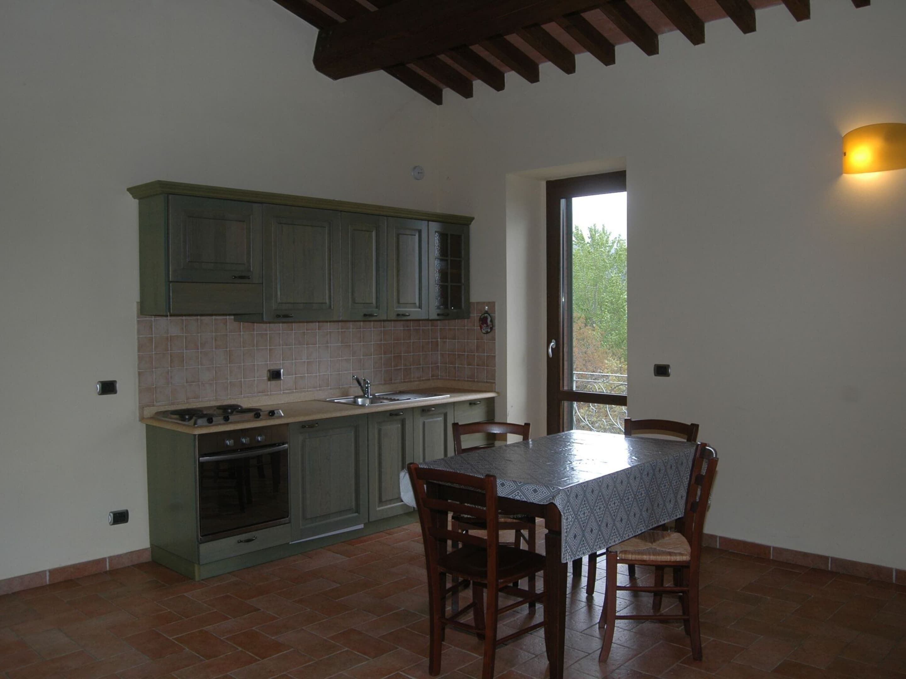 Une cuisine avec des meubles de rangement verts, un mur recouvert de carreaux, une table à manger avec des chaises et une vue à travers une porte vitrée.