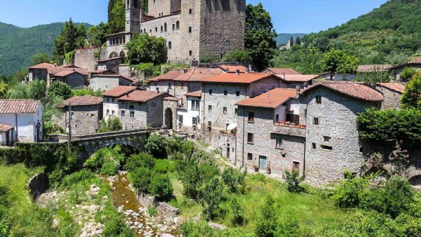 Un castillo de piedra histórico con una torre, rodeado de edificios de piedra tradicionales con techos de tejas rojas, situado en un exuberante valle verde.