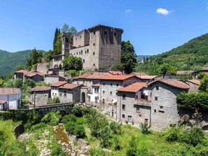 Un château de pierre historique, doté d’une tour, entouré de bâtiments traditionnels en pierre aux toits de tuiles rouges, niché dans une vallée verdoyante.