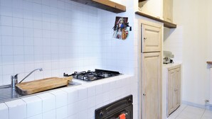 Una cocina con baldosas blancas, estufa a gas, lavamanos, armarios de madera y un cuadro colgado.
