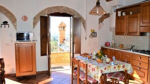 A kitchen with wooden cabinets, a dining table with a floral tablecloth, and a view through an open doorway to a tower. - Apartment in Tuscany With Stunning Views (Asciano)