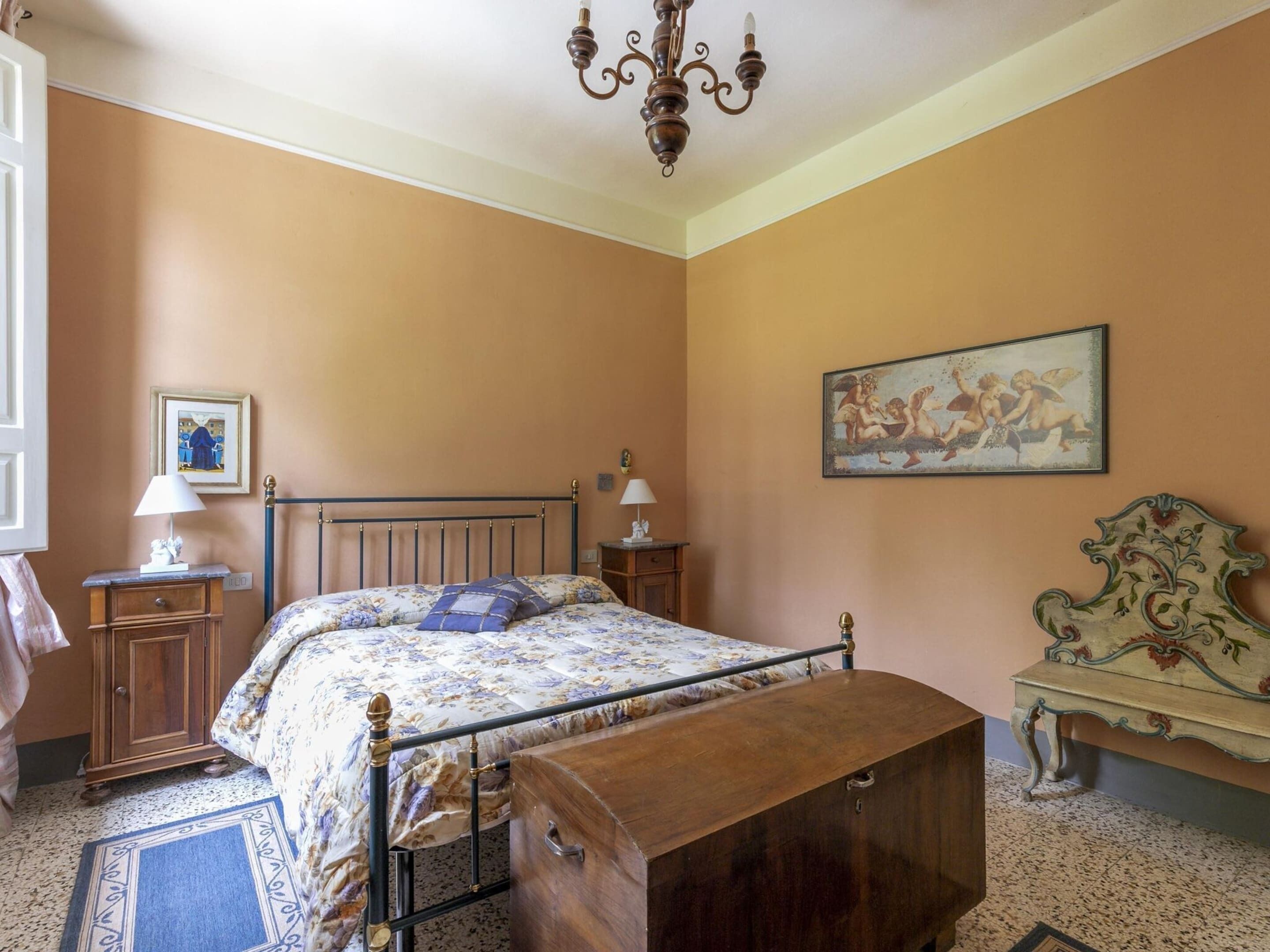 Un dormitorio con cama, mesita de noche, cómoda, silla, candelabro y un cuadro en la pared.