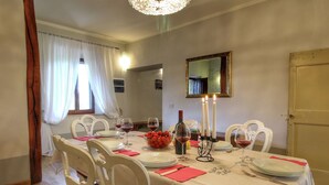 A dining room with a table set for four, white chairs, a chandelier, and a mirror on the wall. - Modern Vacation Home (Pienza)