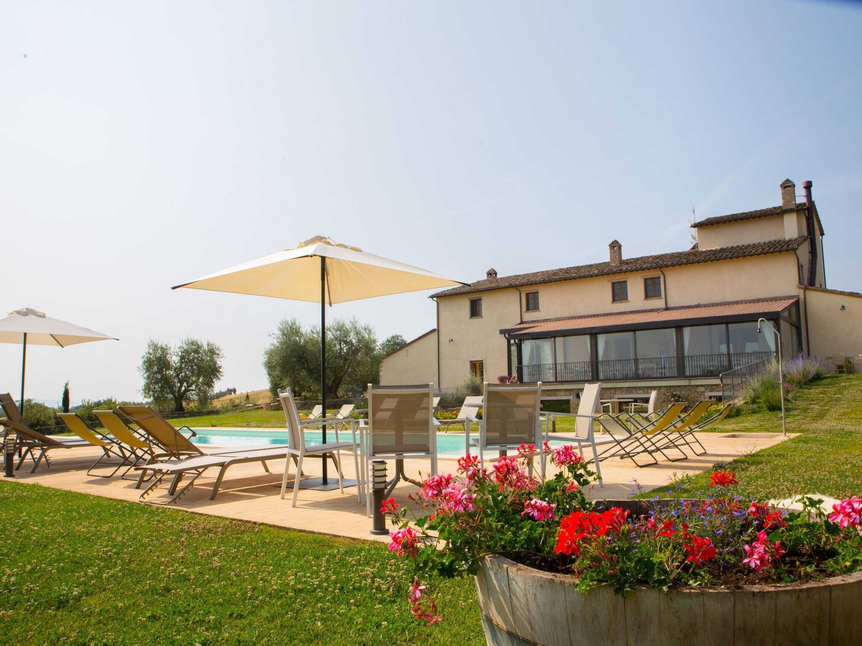 Ein Poolbereich mit Liegestühlen und Sonnenschirmen, ein Haus mit Balkon und ein Blumenbeet.