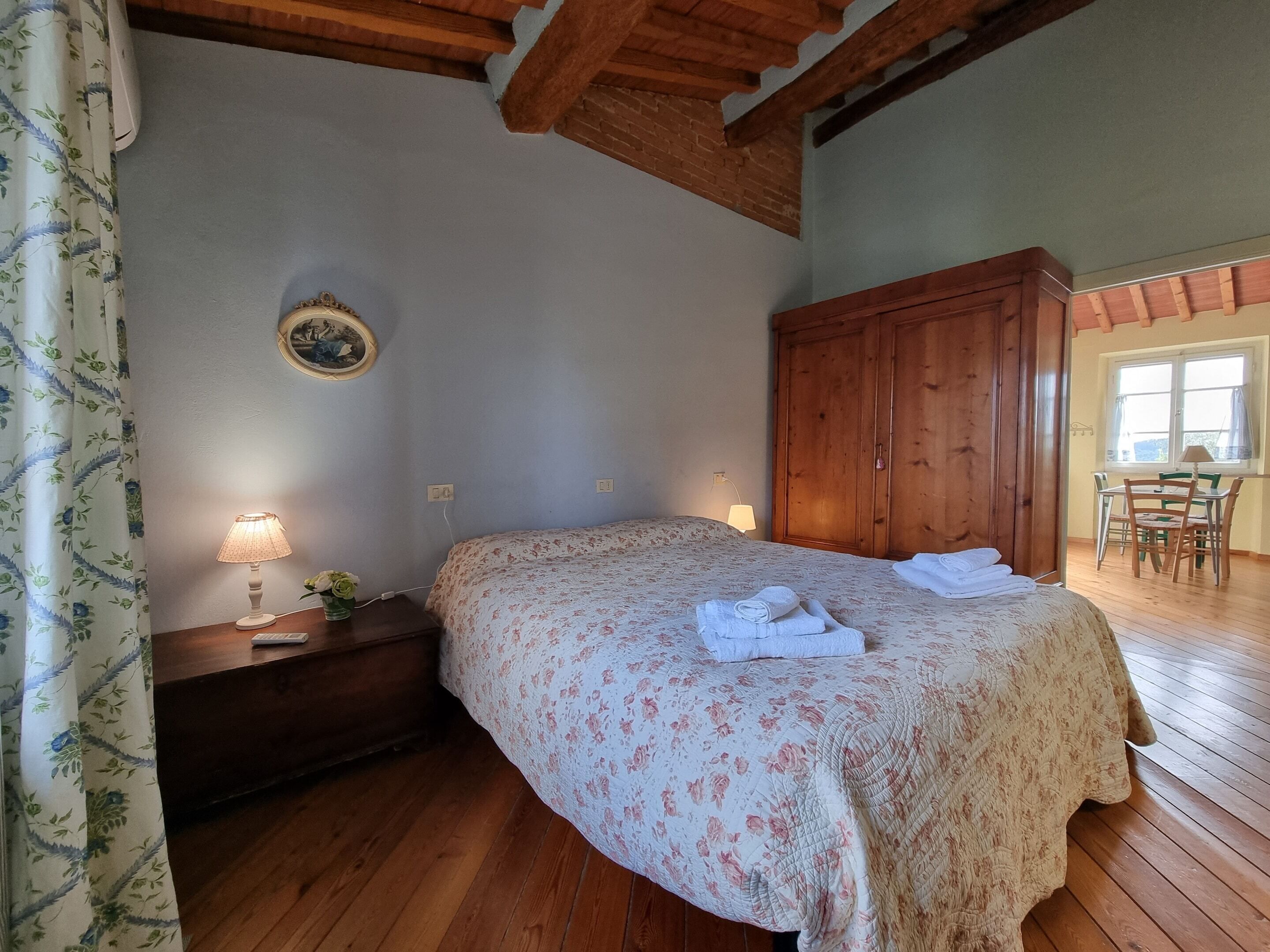 Un dormitorio con una cama de madera, una mesita de noche con lámpara, un armario grande y un área de comedor con sillas y una mesa.