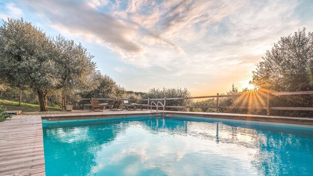 Piscina com deck de madeira e espreguiçadeiras, rodeada por árvores e um pôr do sol ao fundo.