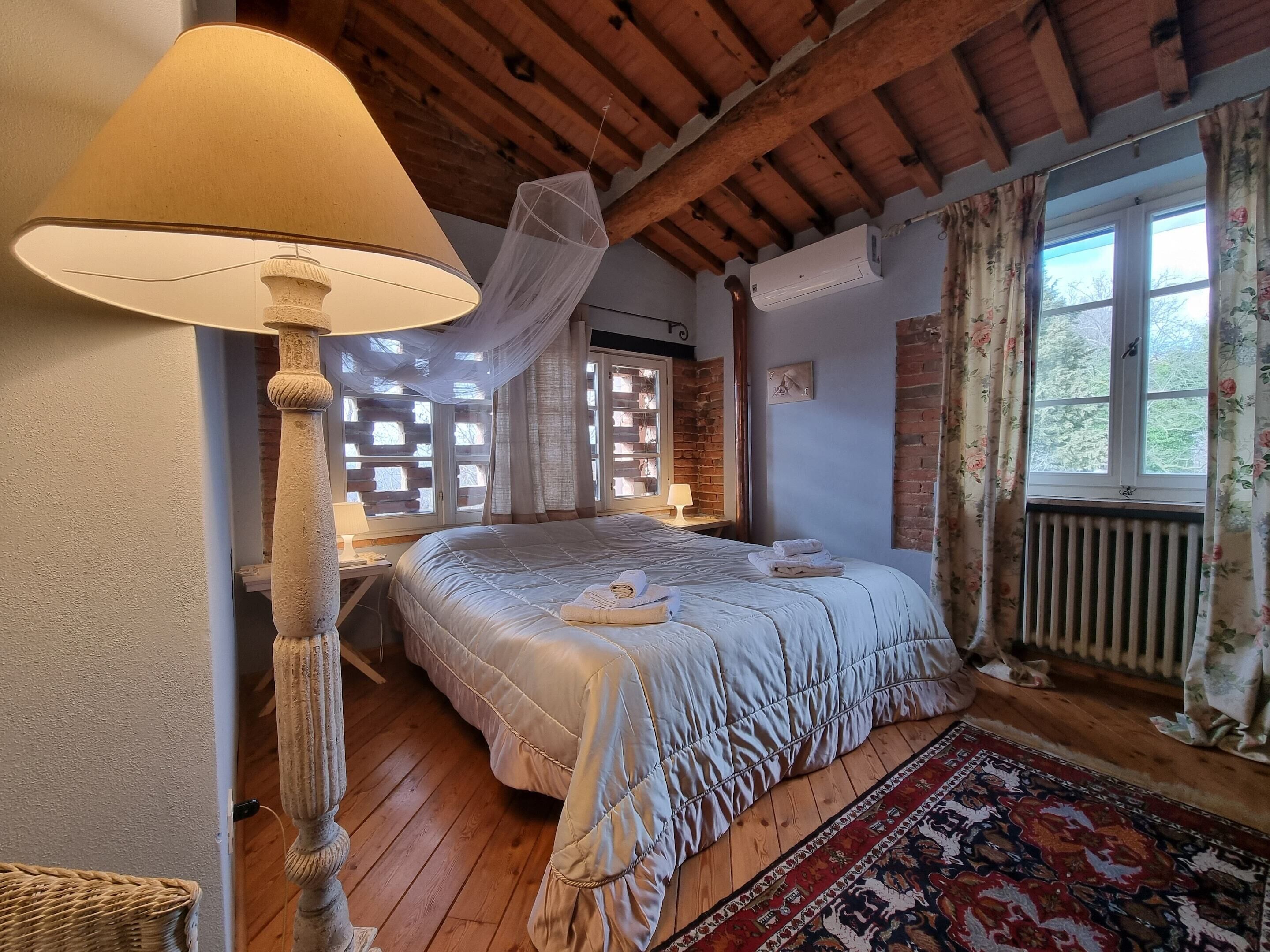 A cozy bedroom with a large bed, a wooden floor, a window with floral curtains, and a wall-mounted air conditioner.