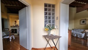 Quarto com piso de madeira, uma pequena mesa com um vaso de flores, uma janela com padrão quadriculado e um sofá.
