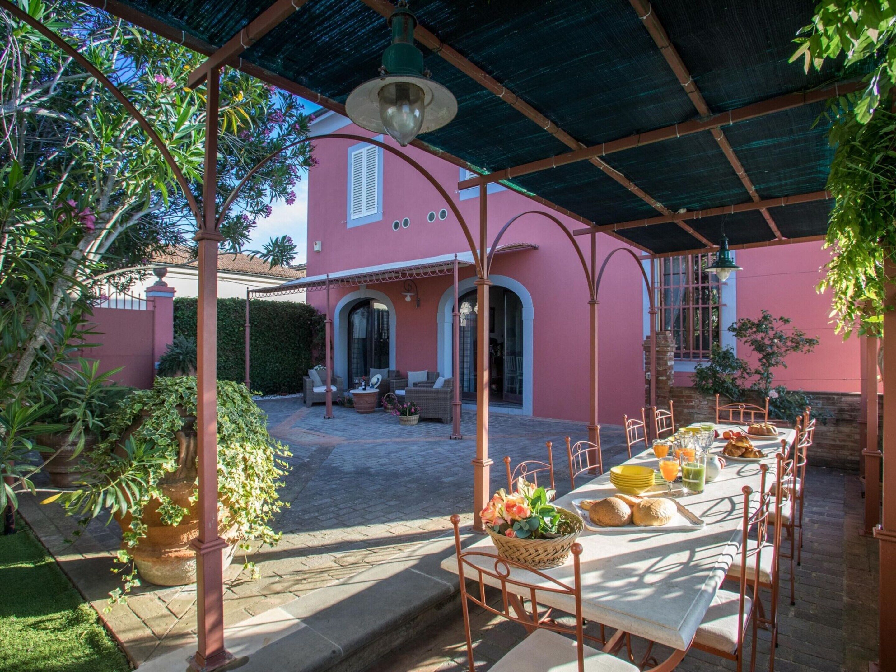 A covered outdoor dining area with tables and chairs, a pergola structure, and a pink building in the background.