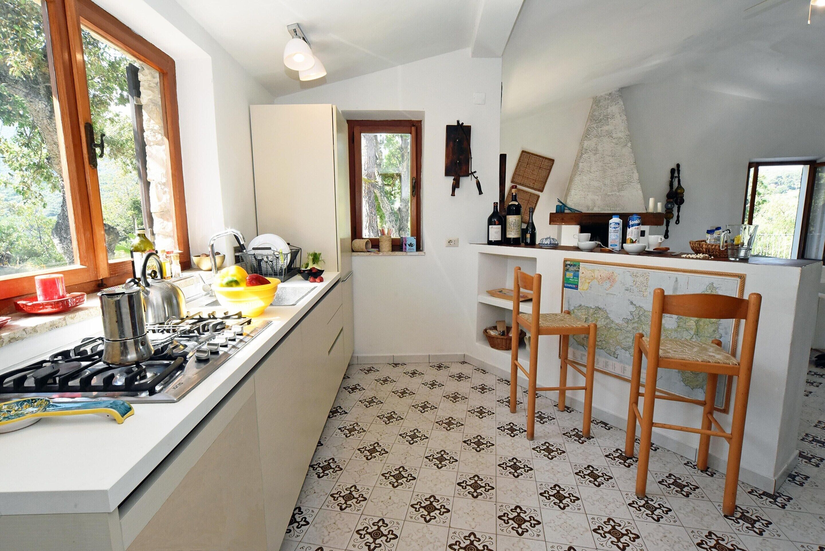 A kitchen with a gas stove, sink, and wooden chairs. There is a window with a view of trees, and a map on the wall.
