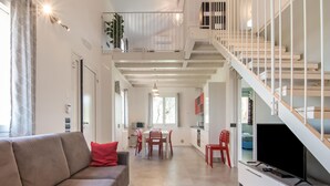 Ein modernes Wohnzimmer mit grauem Sofa, roten Kissen, einem roten Teppich, einem Essbereich mit roten Stühlen, einer Küche mit weißen Schränken und einer Treppe mit weißen Geländern.
