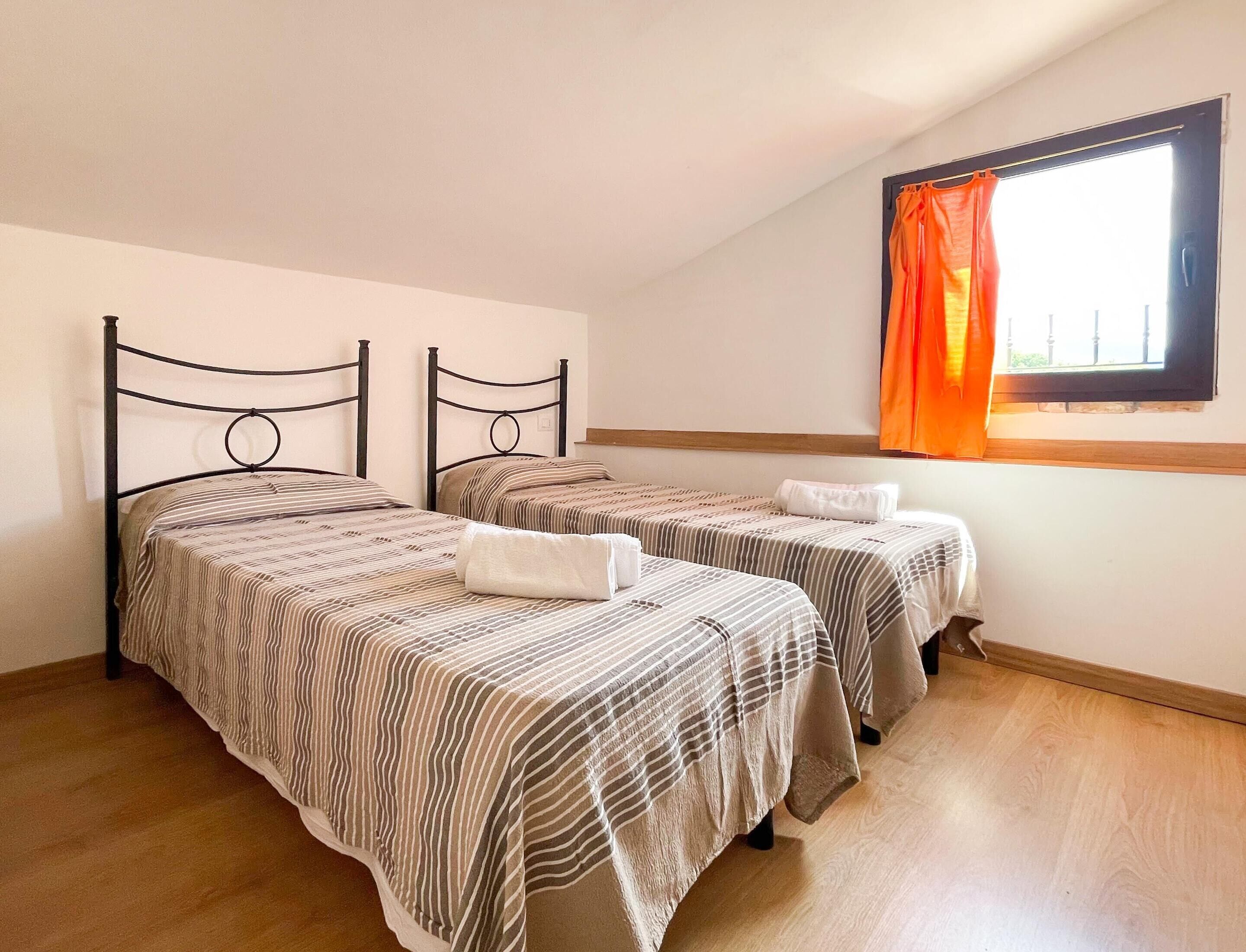 A bedroom with two single beds, a window with orange curtains, and a wooden floor.