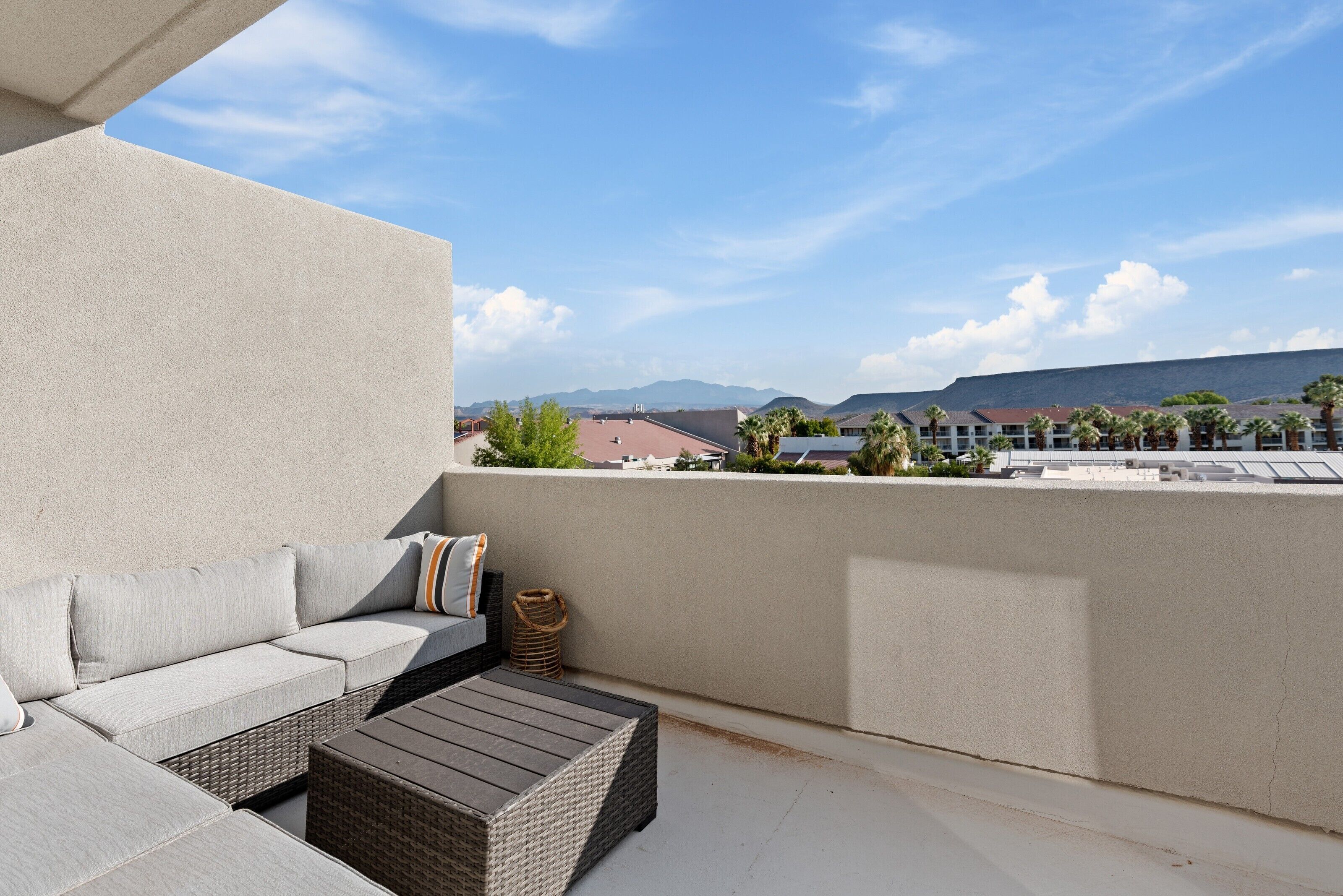 A balcony with a sofa and a table, overlooking a cityscape with buildings and mountains.