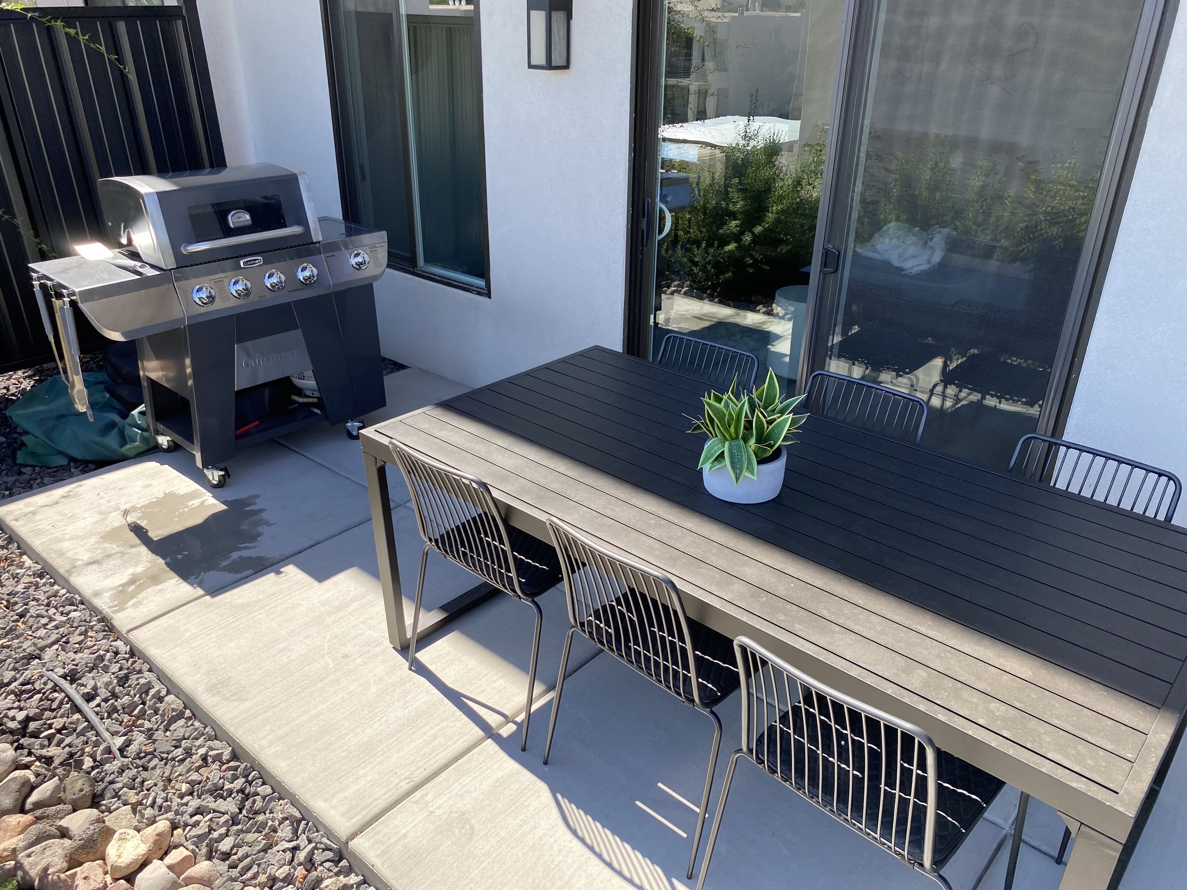An outdoor patio with a wooden table, metal chairs, a grill, and a potted plant.