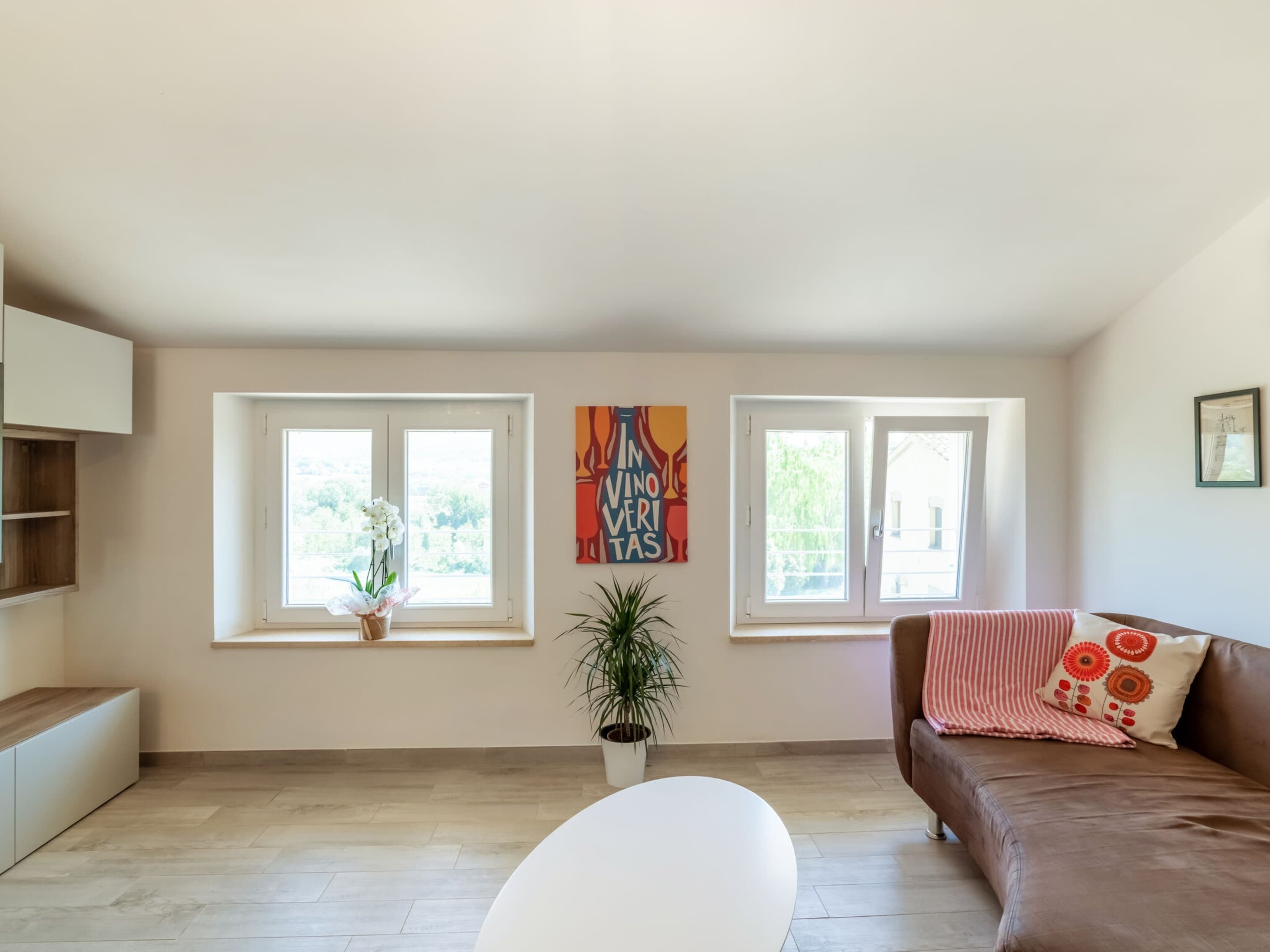 A spacious living room with a brown sofa, a white armchair, and a coffee table. There are two windows with potted plants on the windowsill.