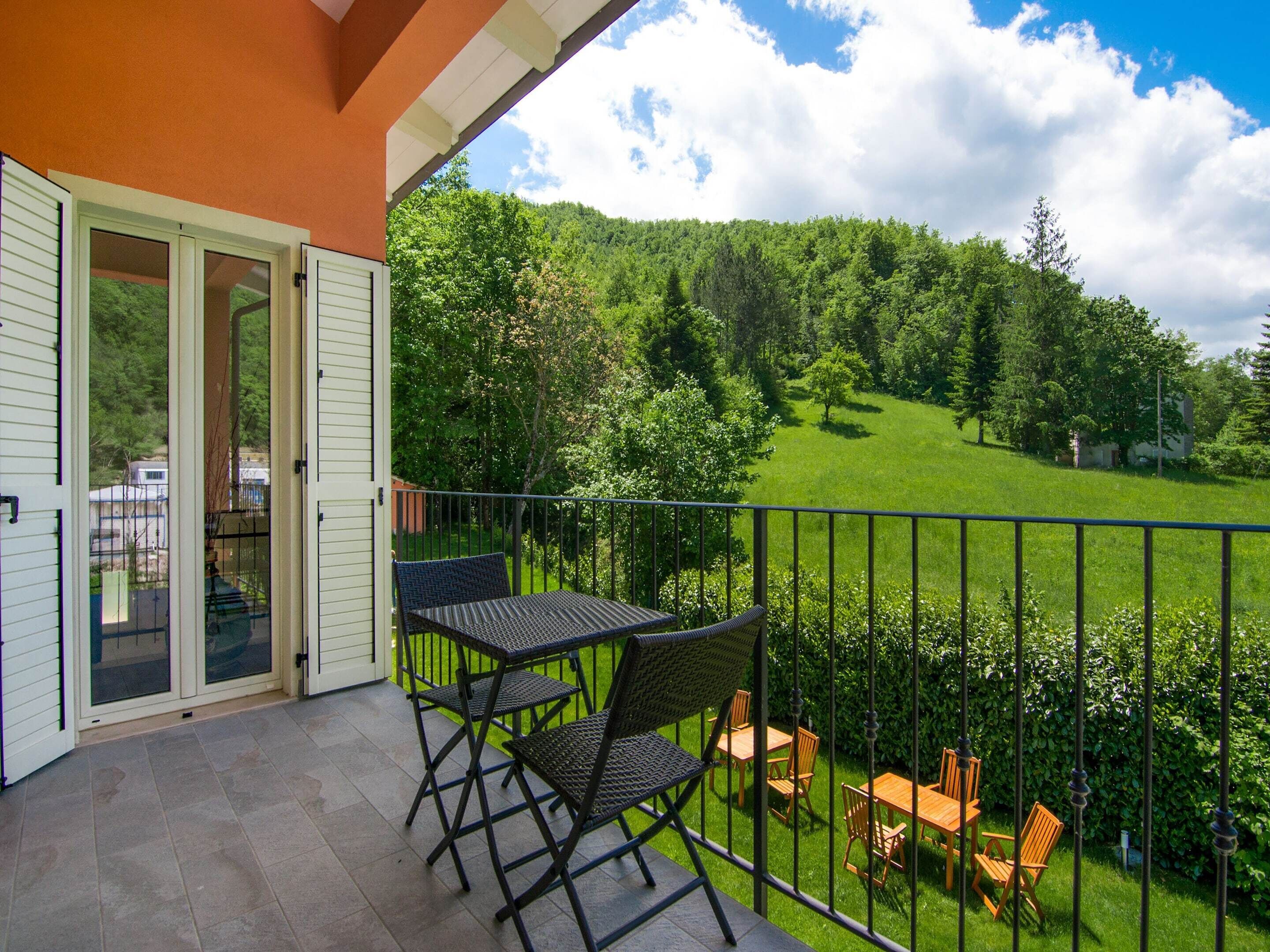 A balcony with a table and chairs, overlooking a green landscape with trees and a hill.