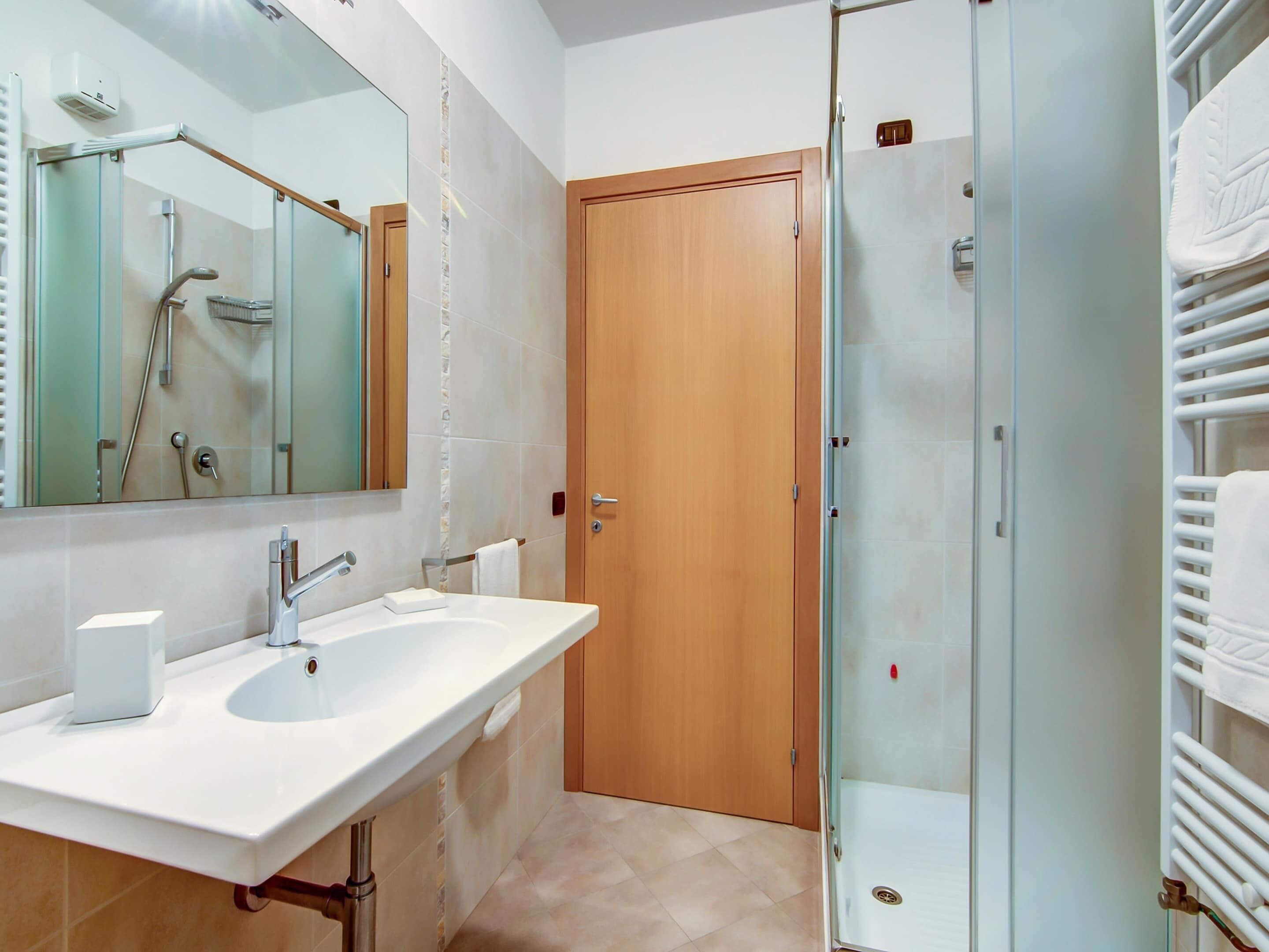 Un baño moderno con cabina de ducha de vidrio, puerta de madera y lavamanos blanco con espejo arriba.