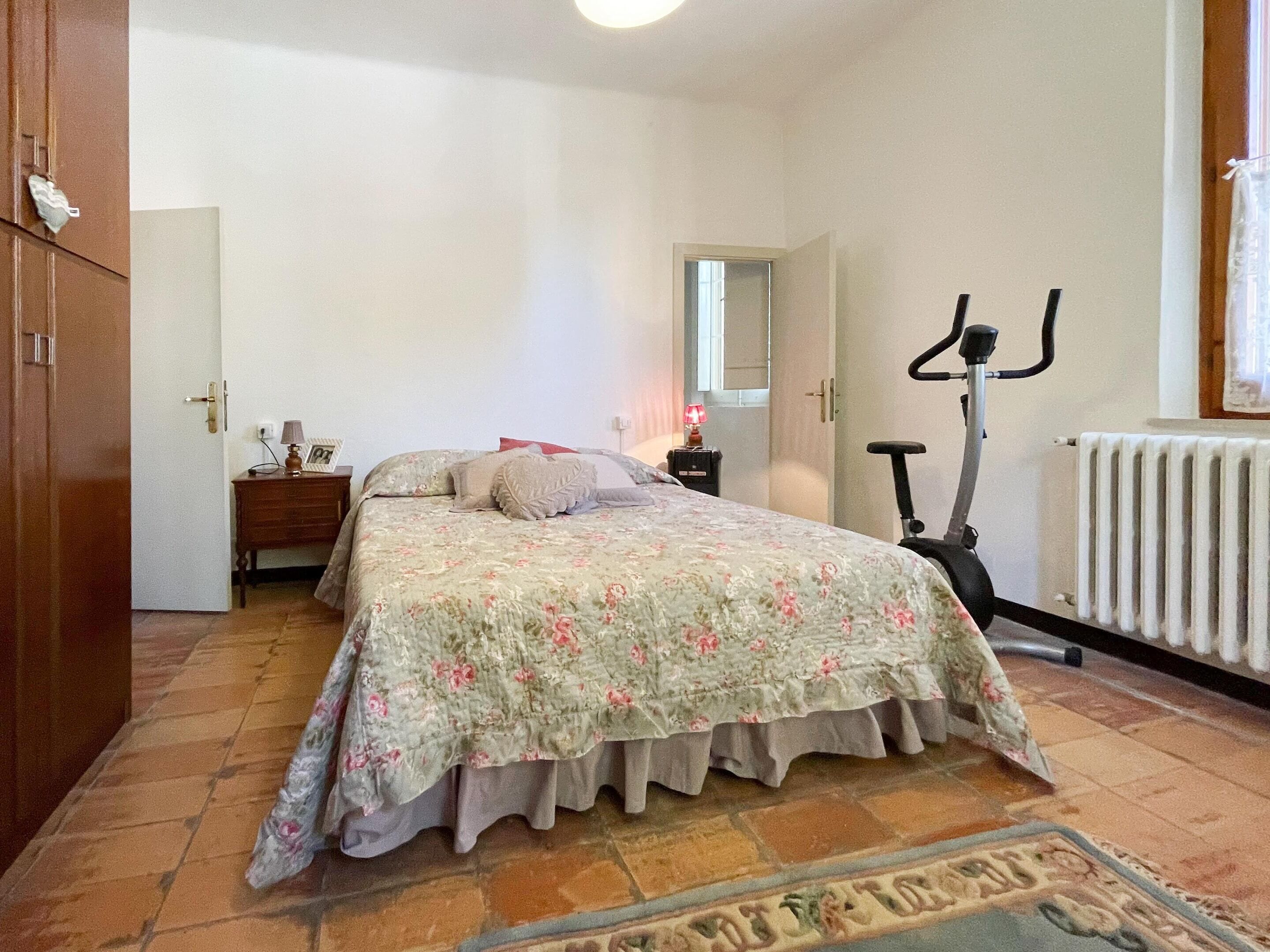 Quarto com cama, penteadeira, espelho, radiador e bicicleta ergométrica.