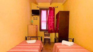 A small, narrow room with two single beds, a wooden wardrobe, a chair, and a television on the wall.