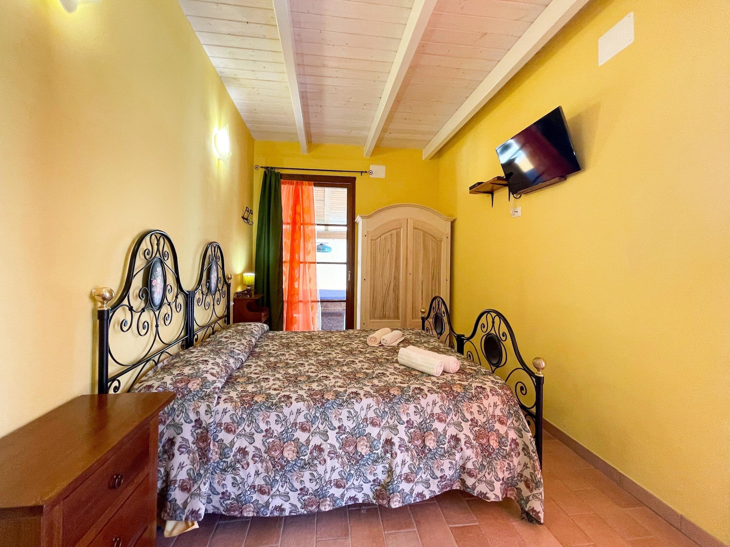 A bedroom with a bed, a wooden dresser, a television mounted on the wall, and a window with curtains.