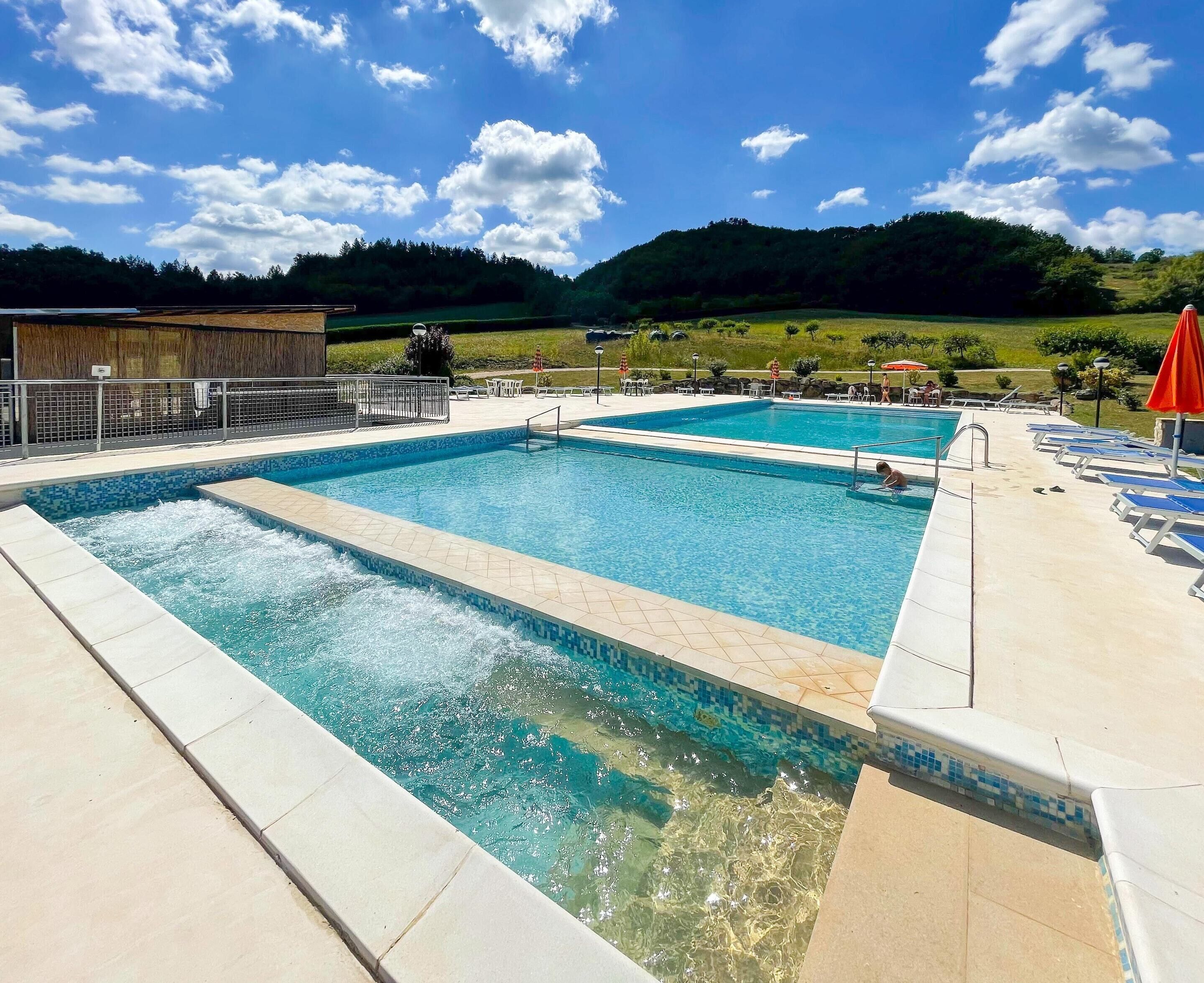 Una piscina con agua azul cristalina, rodeada de sillas de descanso y sombrillas, con colinas verdes y un cielo despejado al fondo.