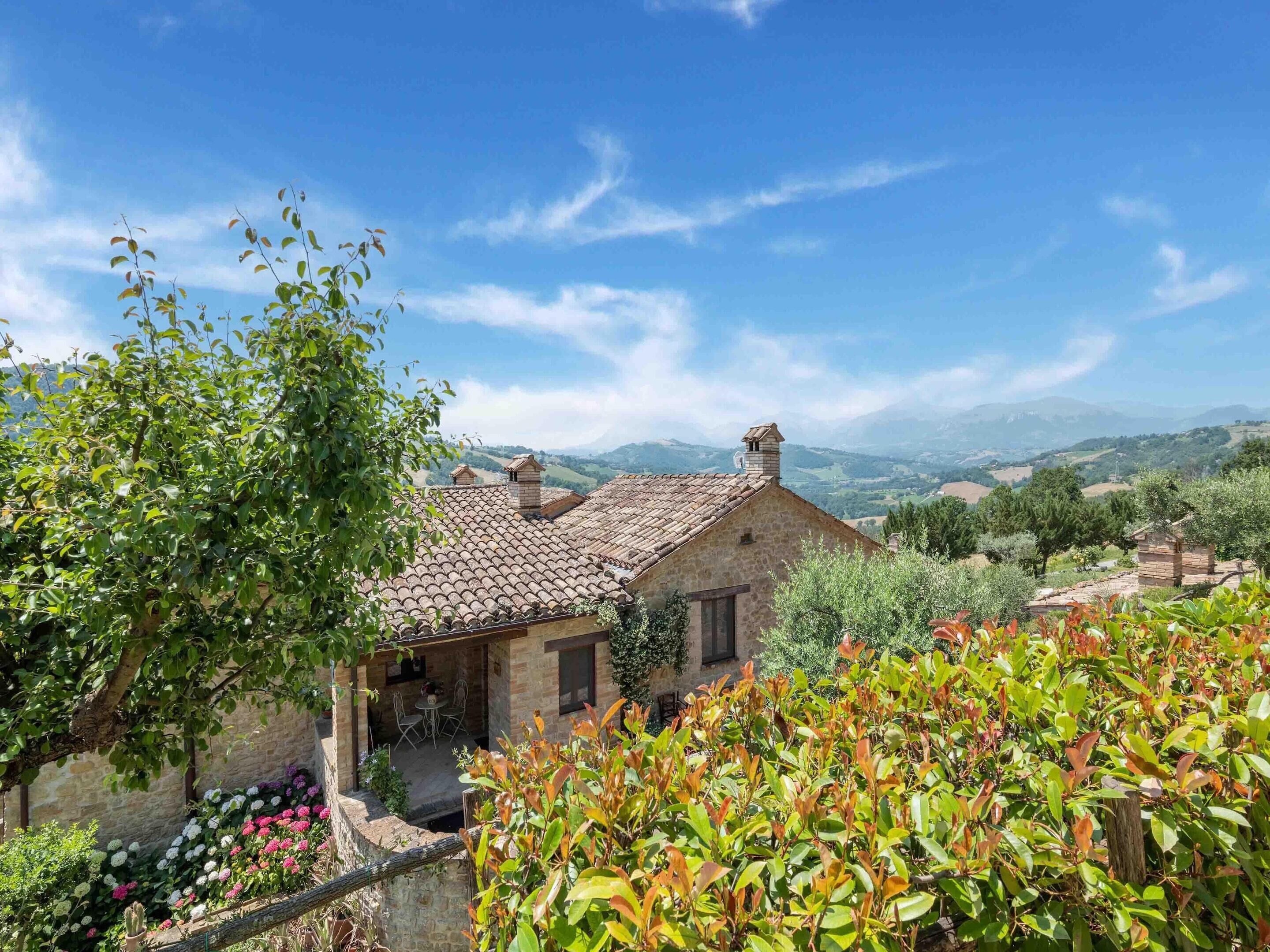 A stone building with a tiled roof, surrounded by lush greenery and a scenic view of distant hills.