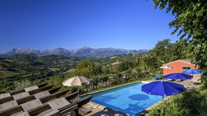 Un espace piscine doté de chaises longues et de parasols, entouré de verdure et avec des montagnes en toile de fond.