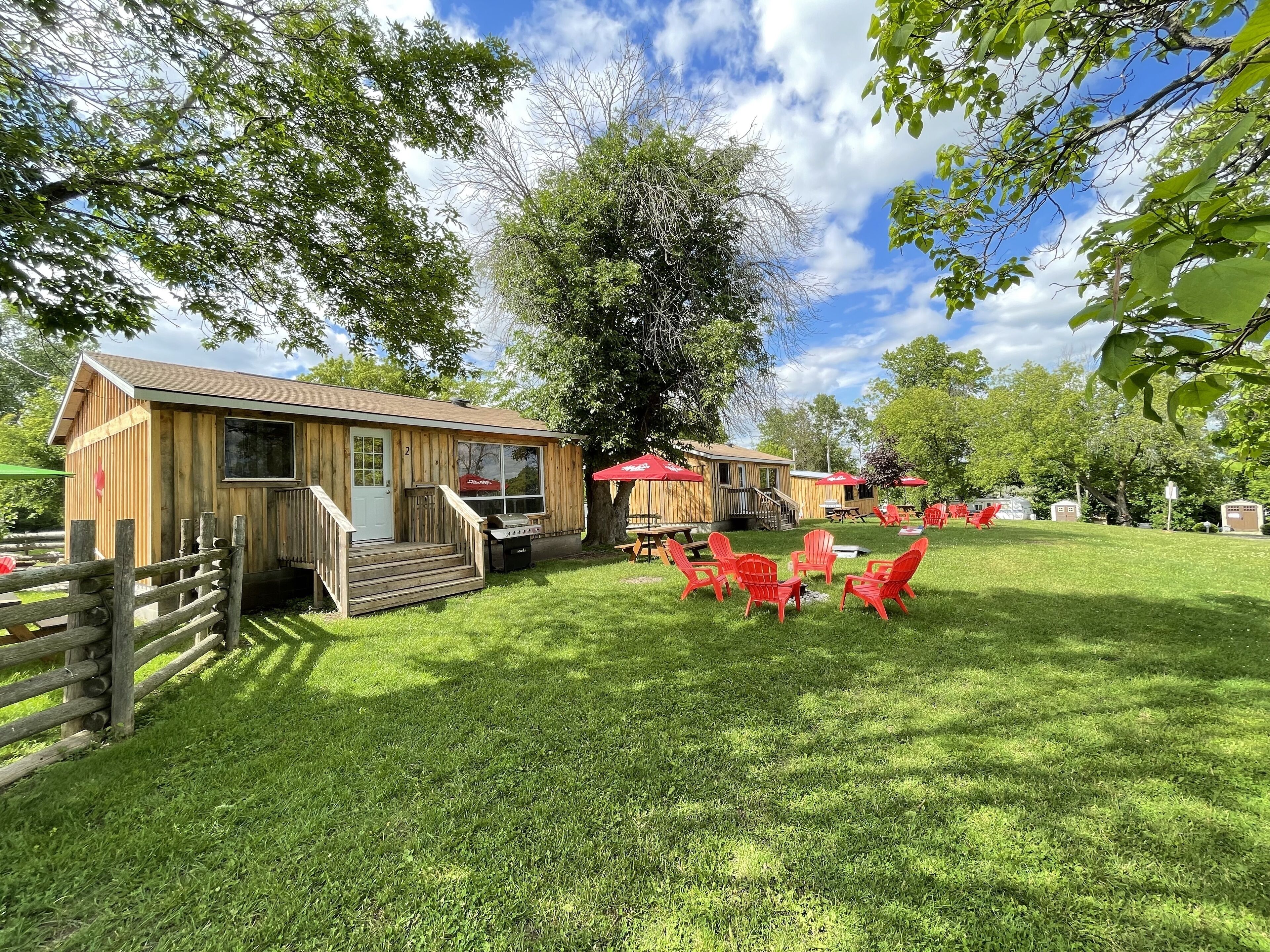 A rustic wooden cabin with a porch and a grill, surrounded by a grassy lawn and red chairs.