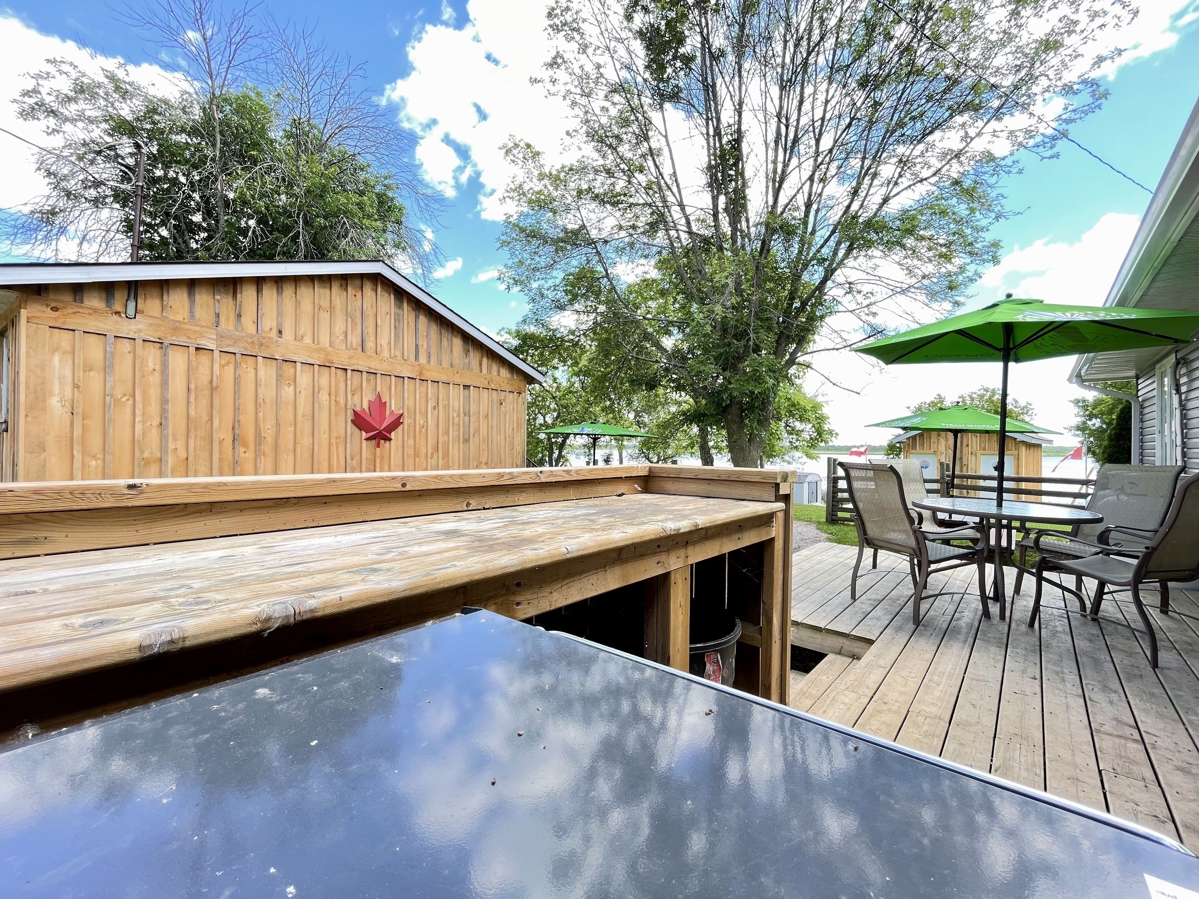 A wooden deck with a table and chairs, a wooden structure with a maple leaf emblem, and a green umbrella.